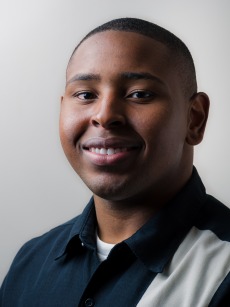 Latimer, wearing a dark shirt with a light stripe, smiles in front of a neutral background.